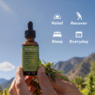 A person holds a dropper bottle of HempLucid Full-spectrum CBD Tincture in Hemp Seed Oil in front of a mountain landscape. The amber glass bottle has a green label and a black dropper cap. with a sun icon and 'relief, a stretching icon and 'recover', a bed icon and 'sleep', and a calendar icon and 'everyday'.